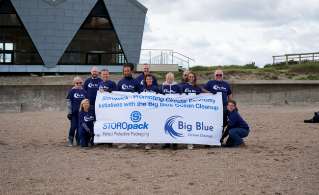 Mehrere Menschen sind zu einem Gruppenfoto am Strand aufgestellt und halten einen Banner des Big Blue Ocean Cleanup.